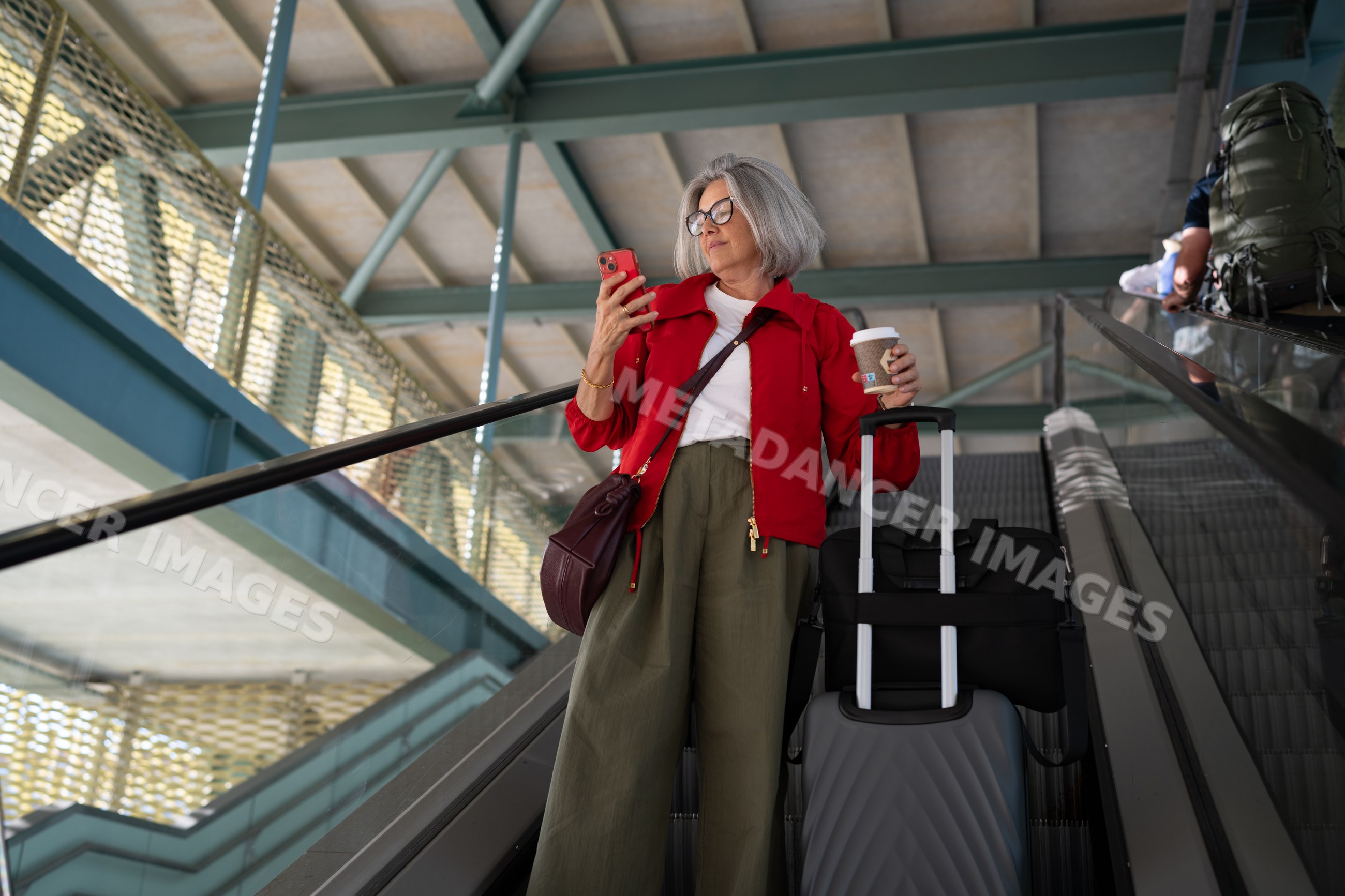 Middle-aged white woman using phone on airport escalator with luggage