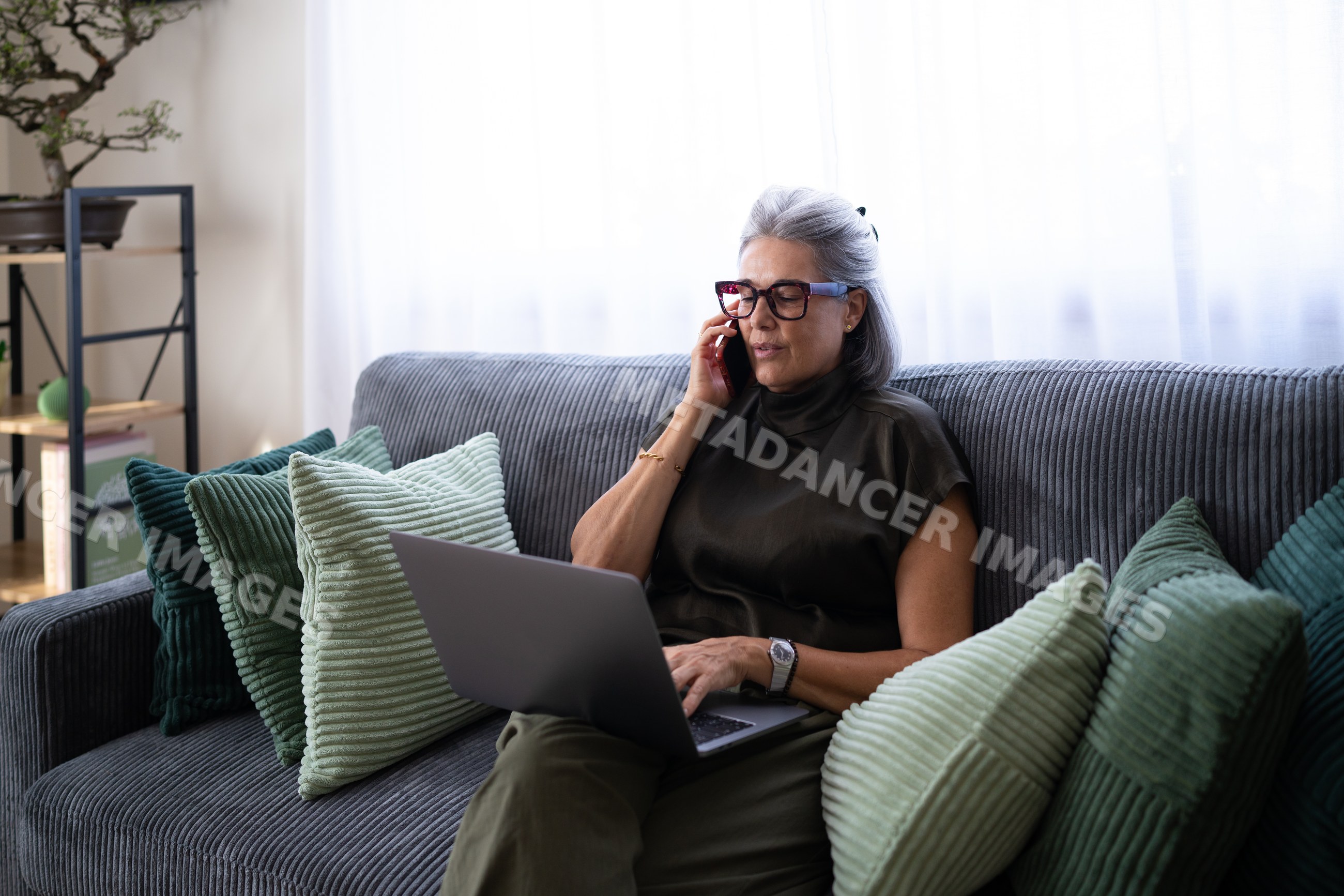 White woman sitting on couch using laptop and talking on phone indoors