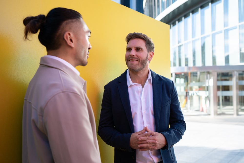 Two white men conversing outdoors near yellow wall and office building