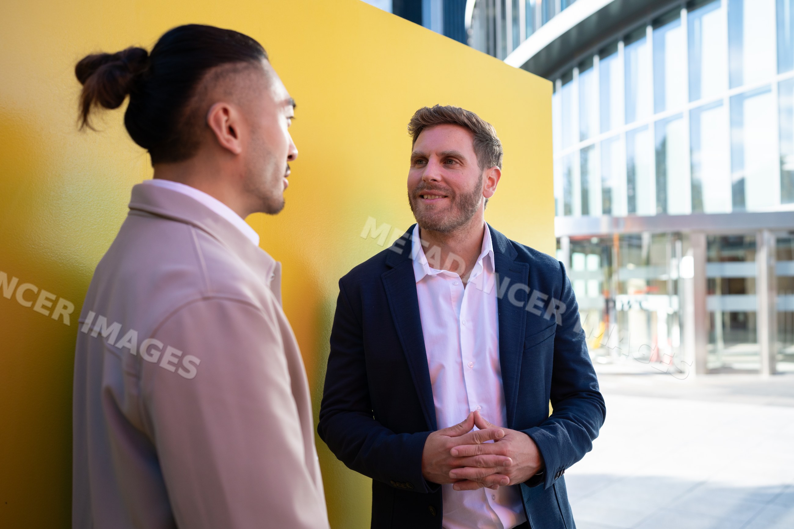 Two white men conversing outdoors near yellow wall and office building