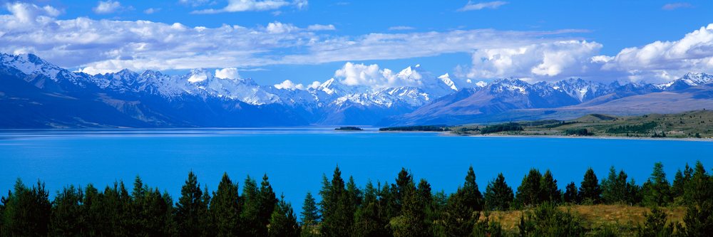 Lake with snow-capped mountains and pine trees under blue sky