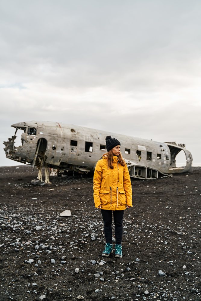 A white woman stands by a crashed airplane wreck on volcanic terrain