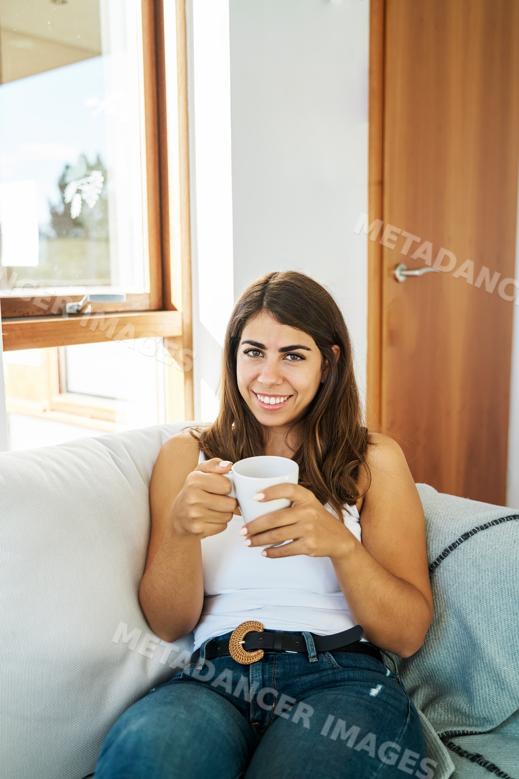 A young white woman sits on a couch holding a cup and smiling