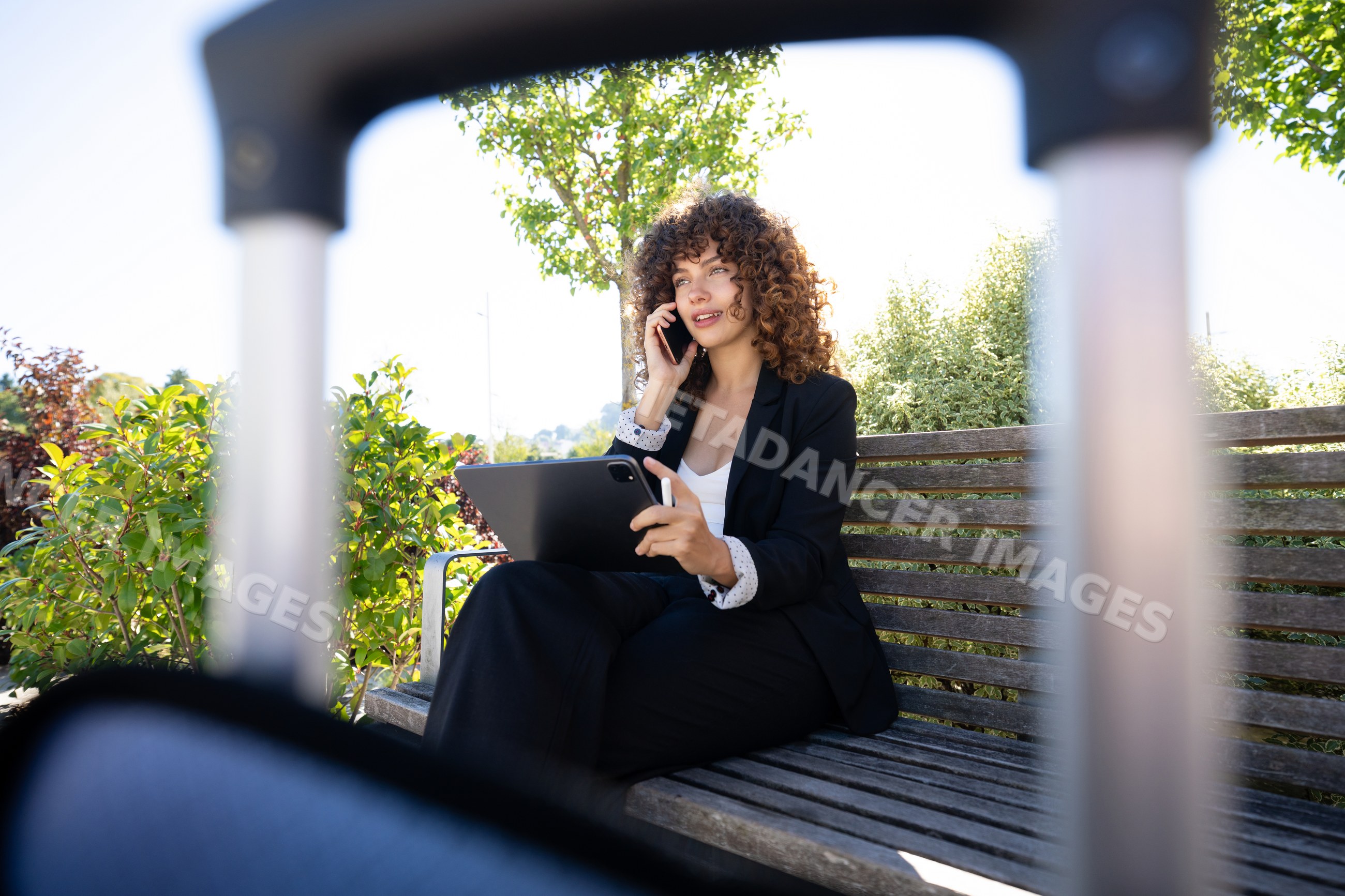 White woman talking on phone and using tablet sitting on outdoor bench