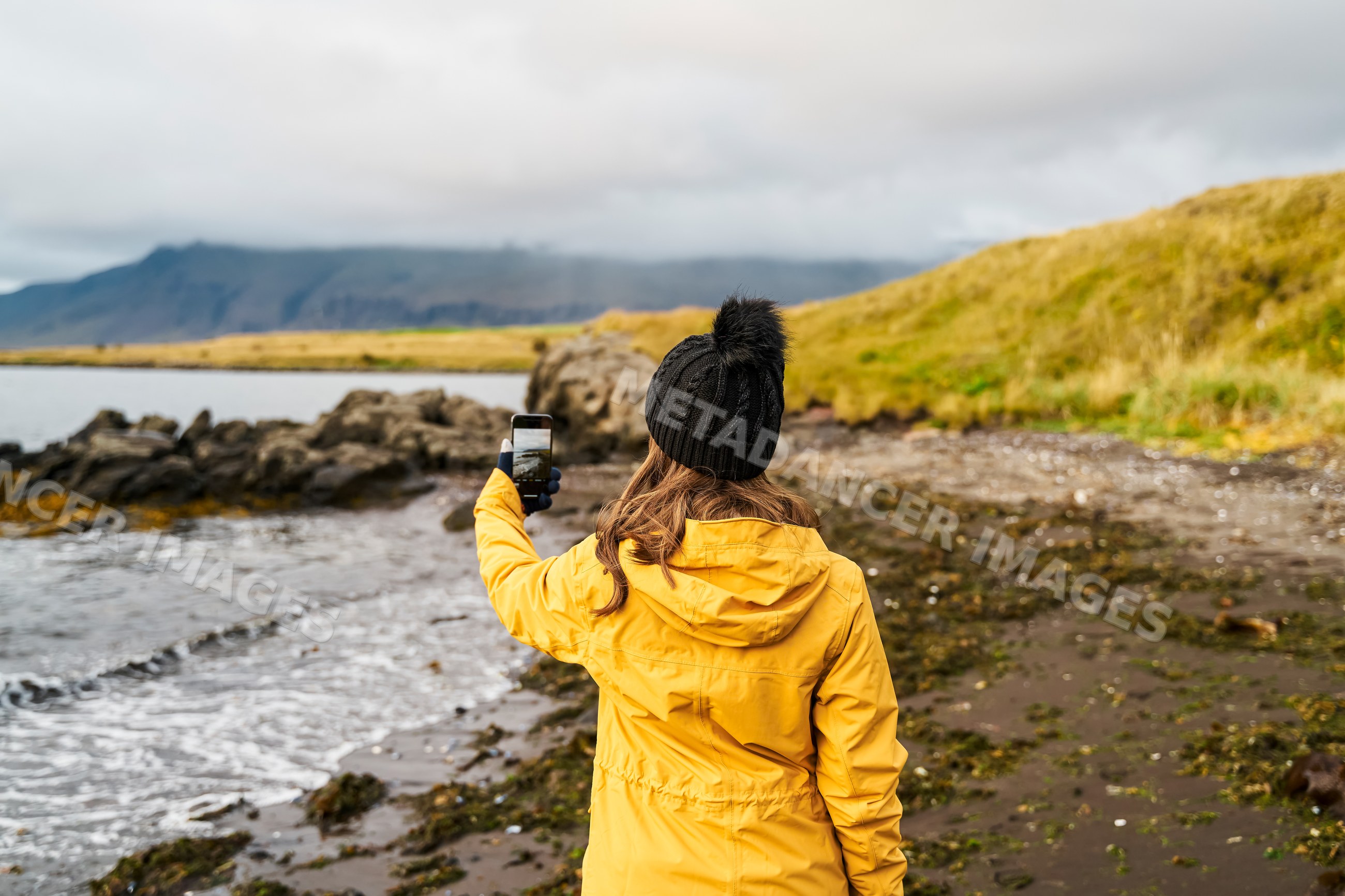 A white woman takes a phone photo on a rocky shore