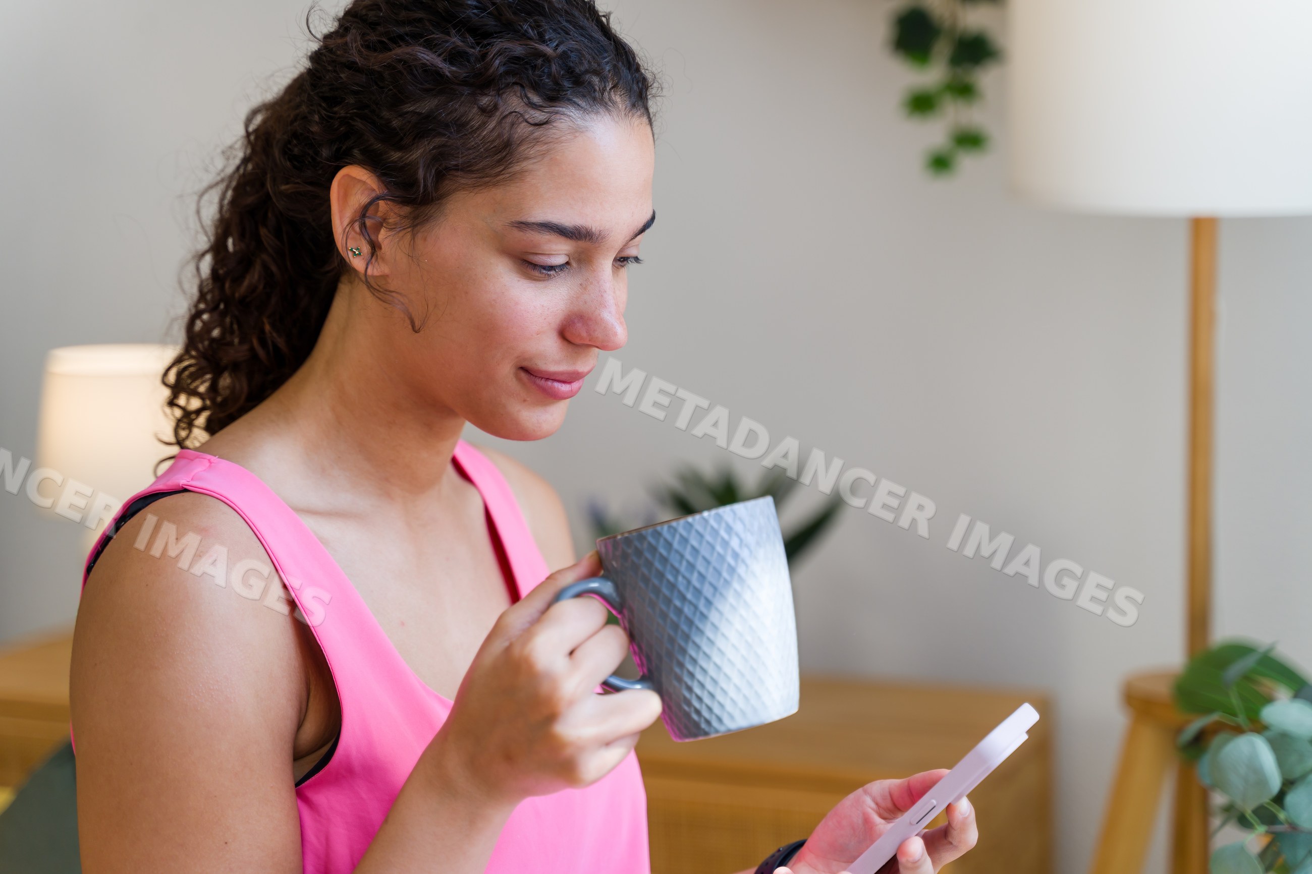 A Hispanic woman in a pink top holds a mug and uses a smartphone