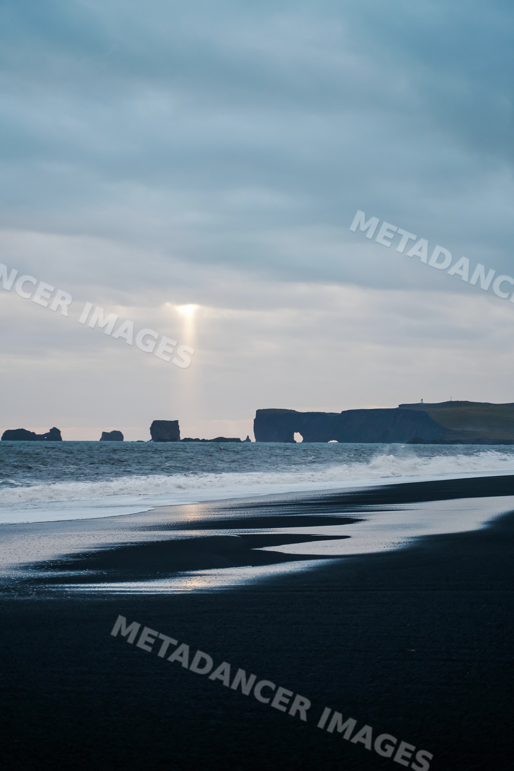 A sunbeam shines through clouds over black sand beach and ocean rocks