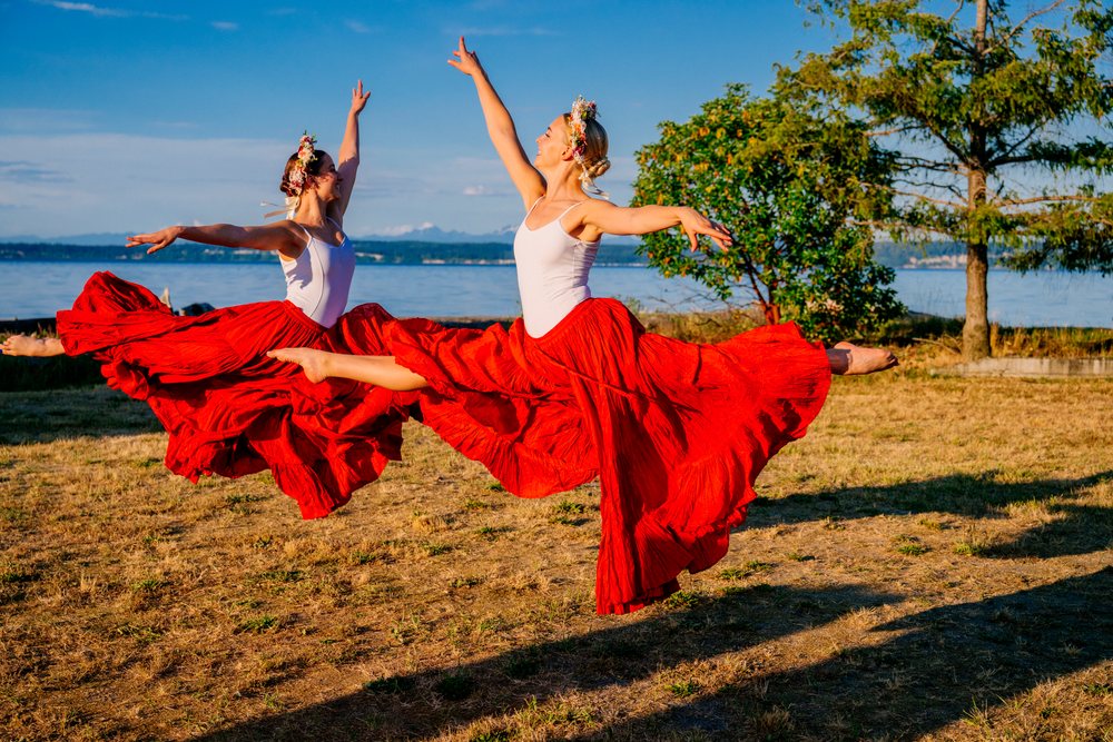 Two white women dancing and leaping outdoors in red skirts near water