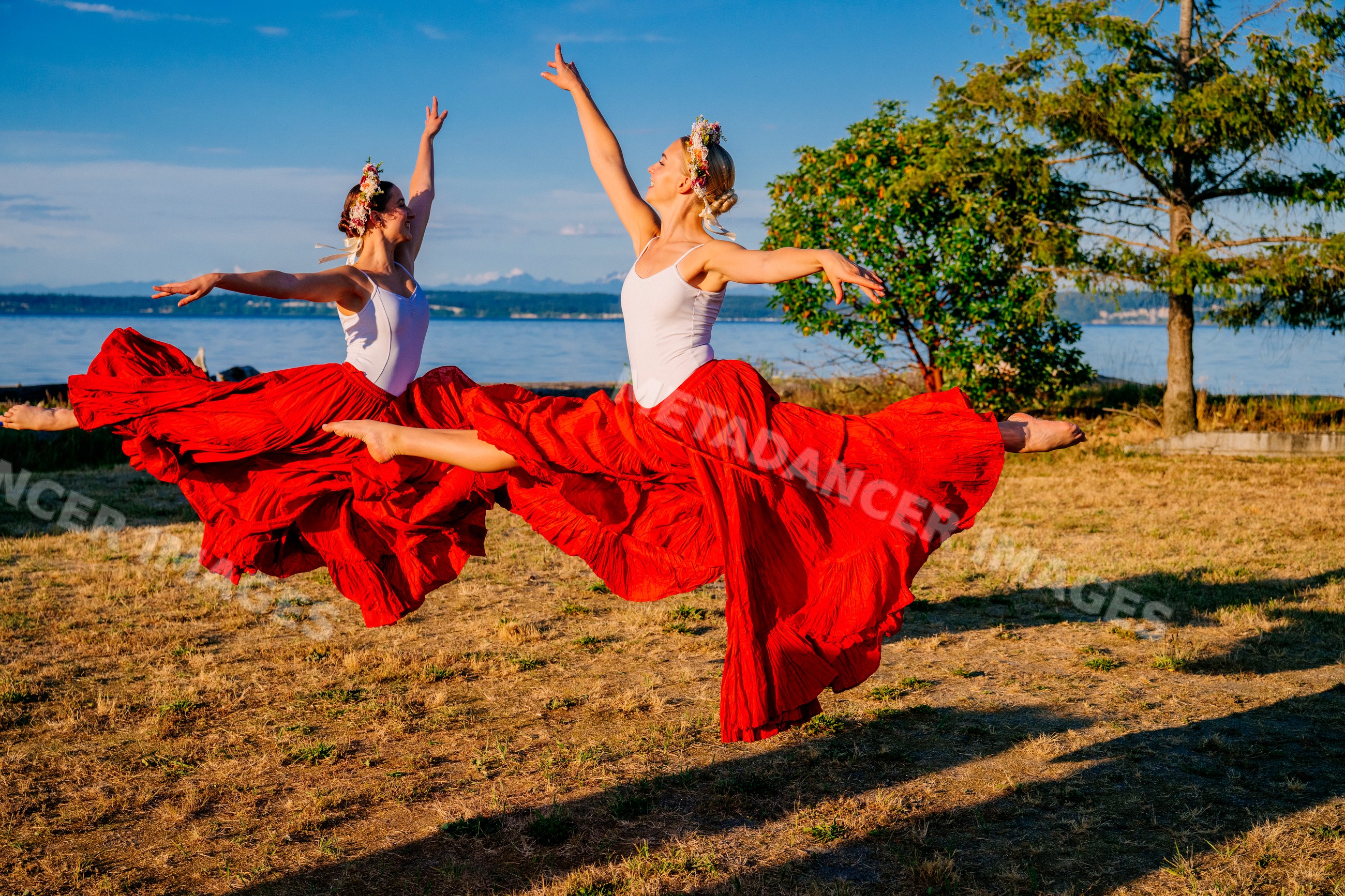 Two white women dancing and leaping outdoors in red skirts near water
