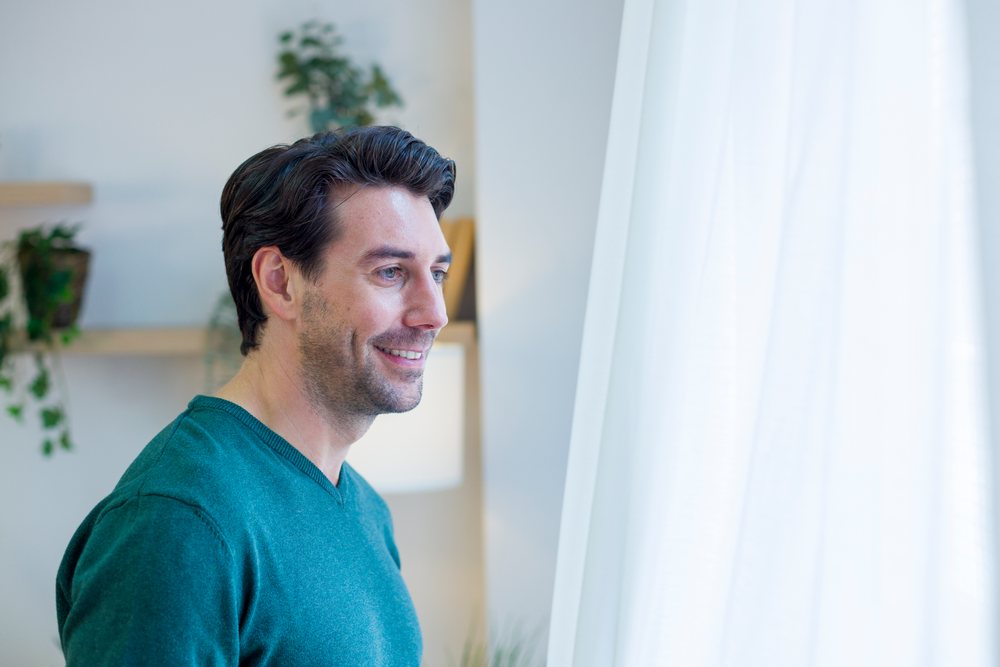 White man smiling and looking out window inside home