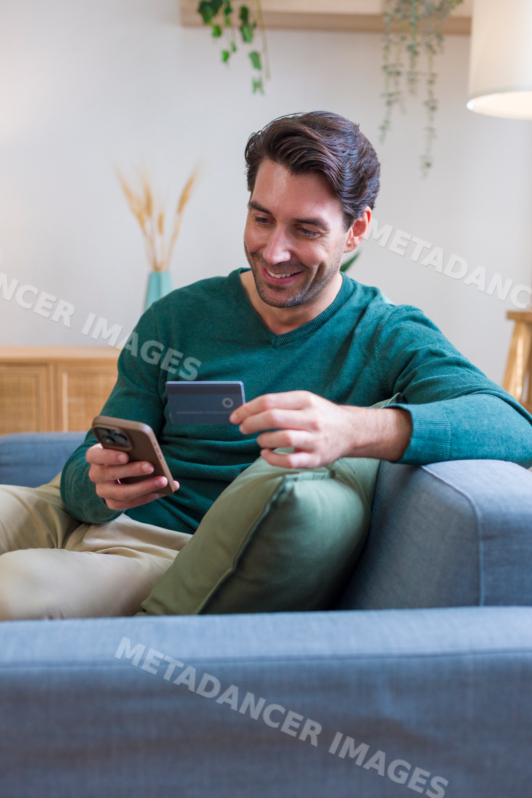 White man holding credit card using smartphone on sofa