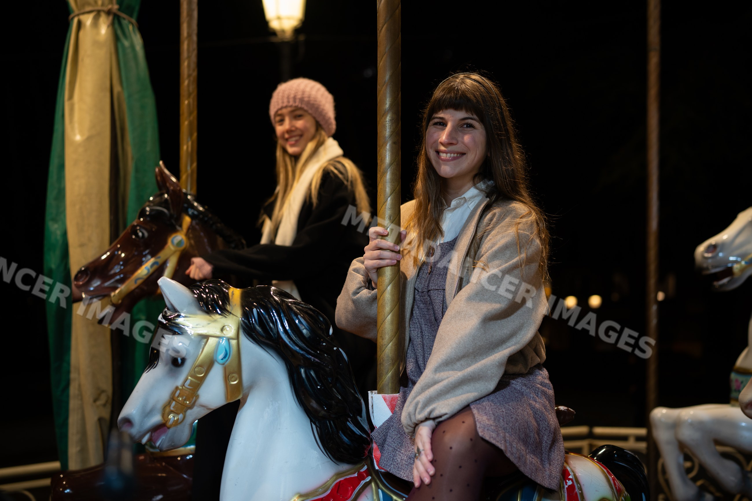 Two white women smiling on merry-go-round carousel horses at night