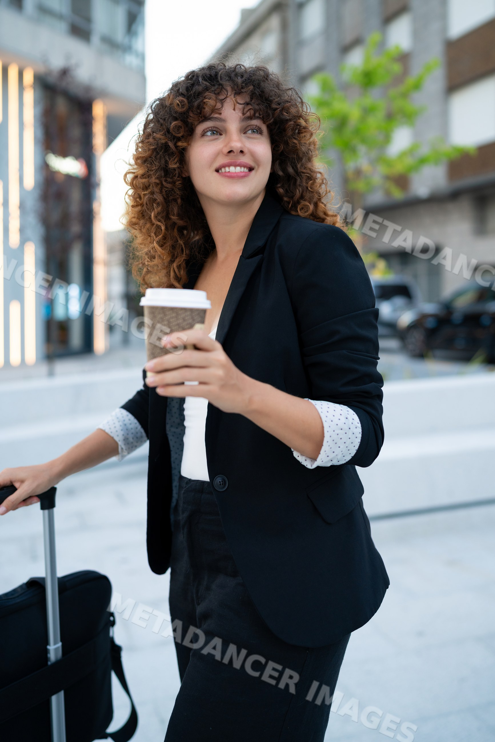 Young white woman holding coffee cup walking with suitcase on urban