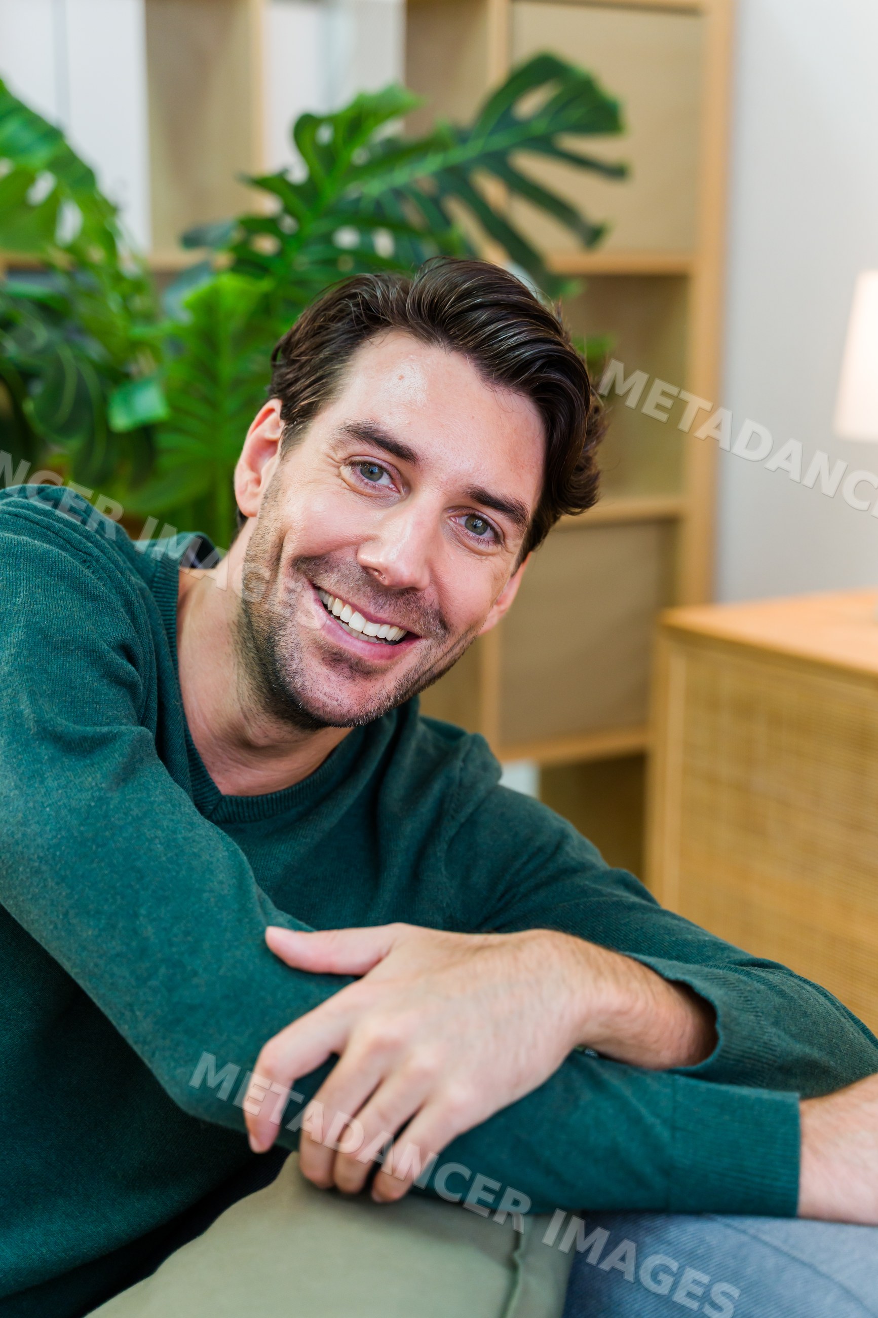 White man smiling and relaxing indoors near houseplants and furniture