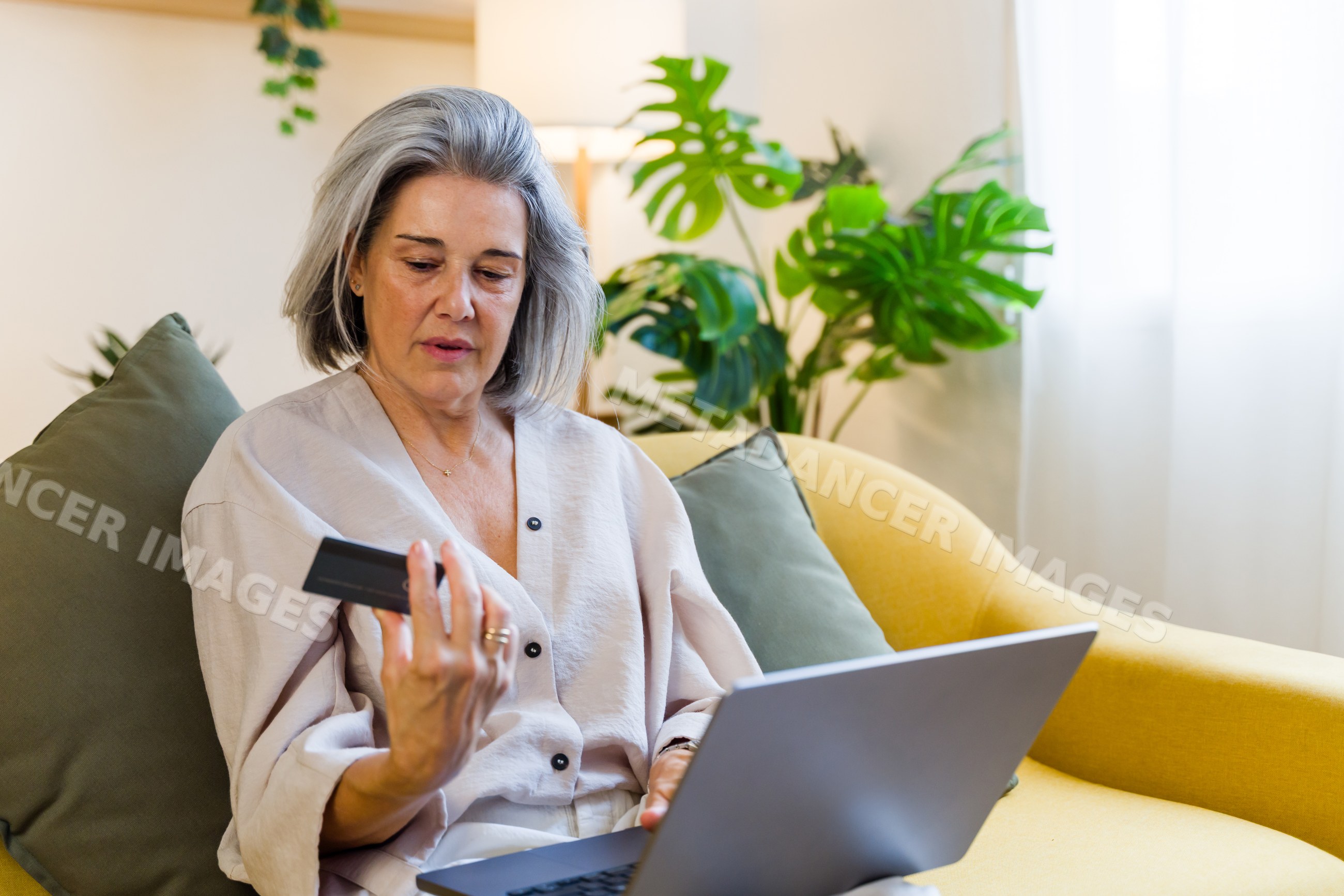 Middle-aged white woman with credit card using laptop on yellow couch