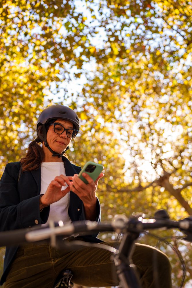 Hispanic woman in helmet using smartphone on bicycle in autumn park