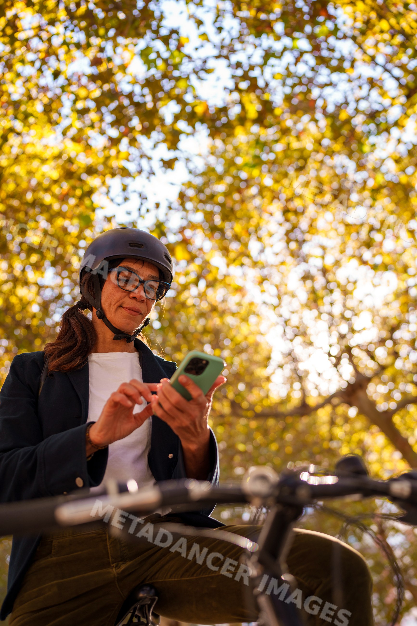 Hispanic woman in helmet using smartphone on bicycle in autumn park