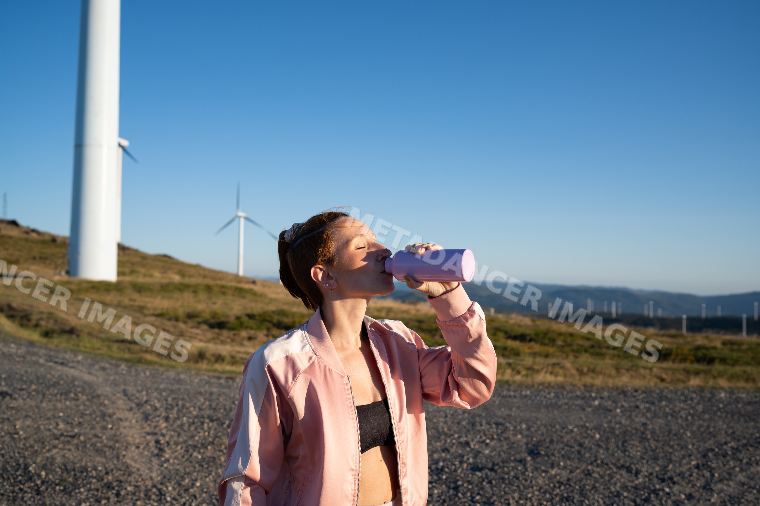 A young white woman drinks water at a wind farm in Galicia, Spain