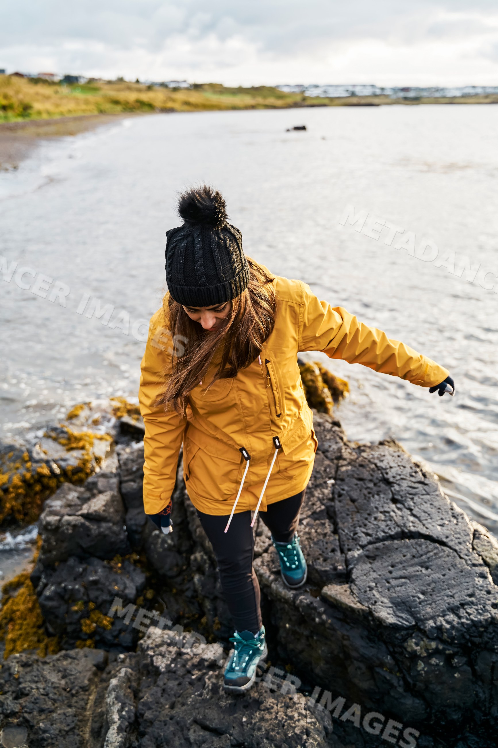 A white woman walks carefully on rocky shore wearing warm clothing