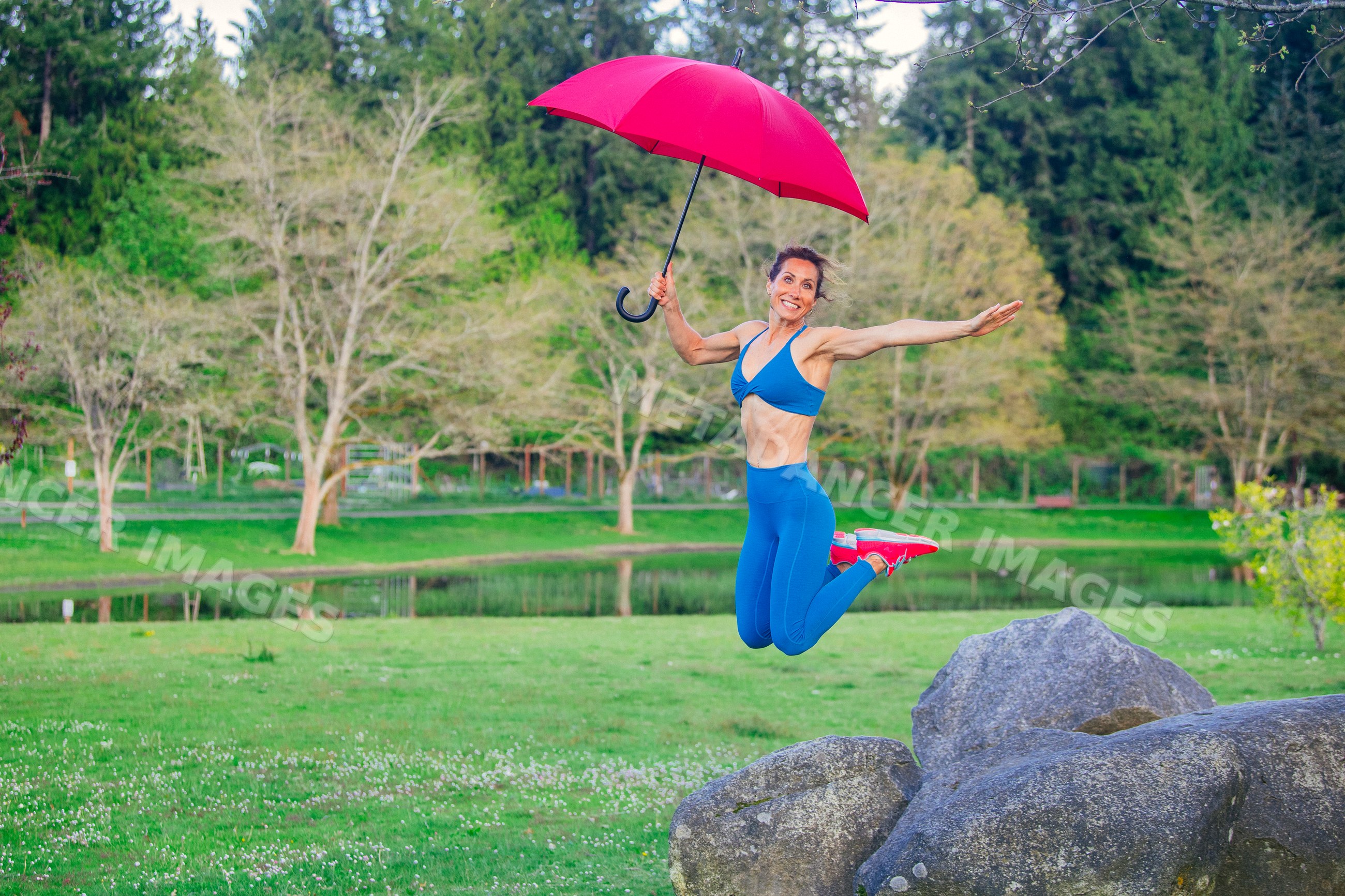 White woman jumping with red umbrella in park with rocks and pond