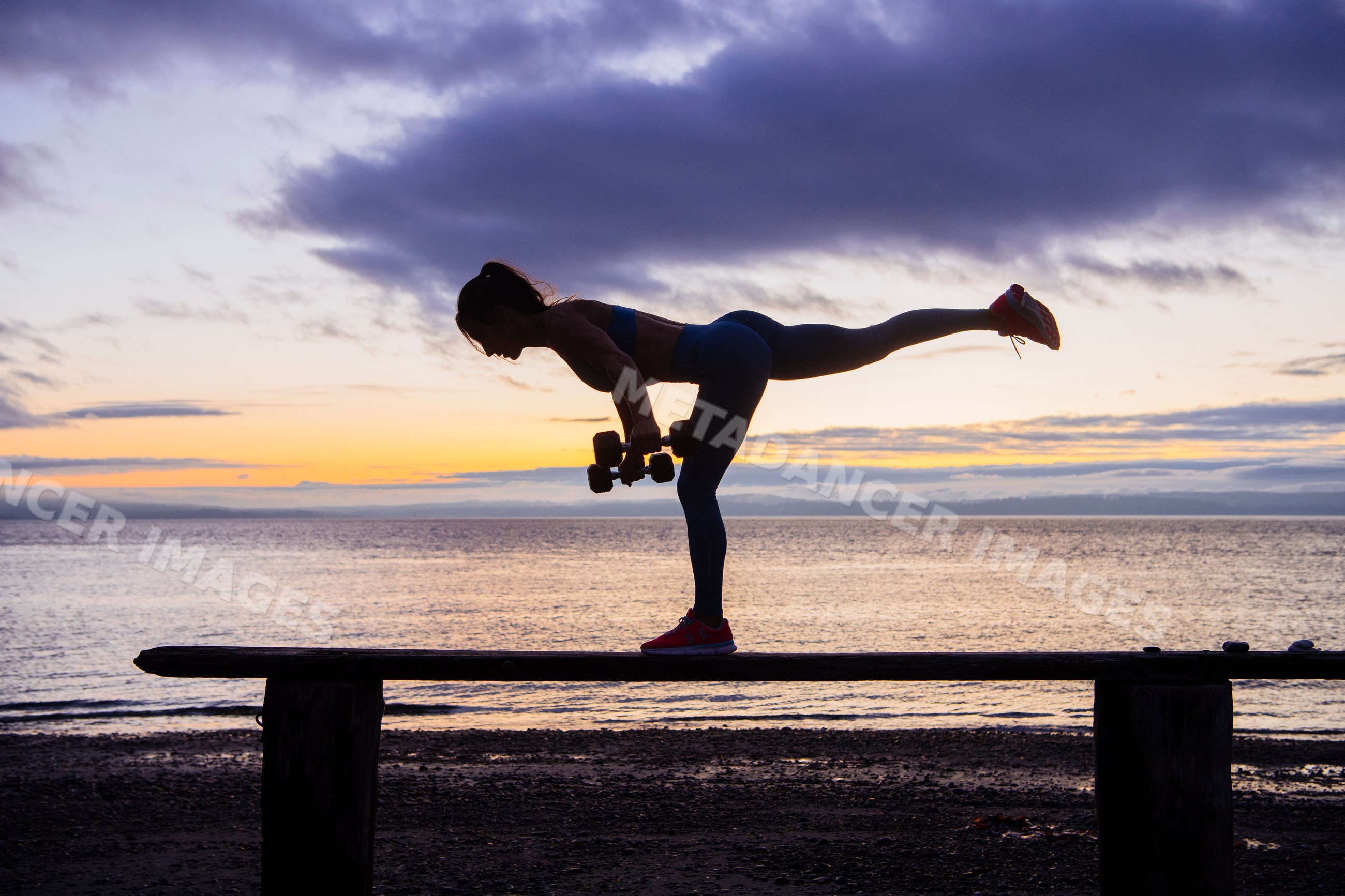 White woman balancing with dumbbells on bench at beach sunset