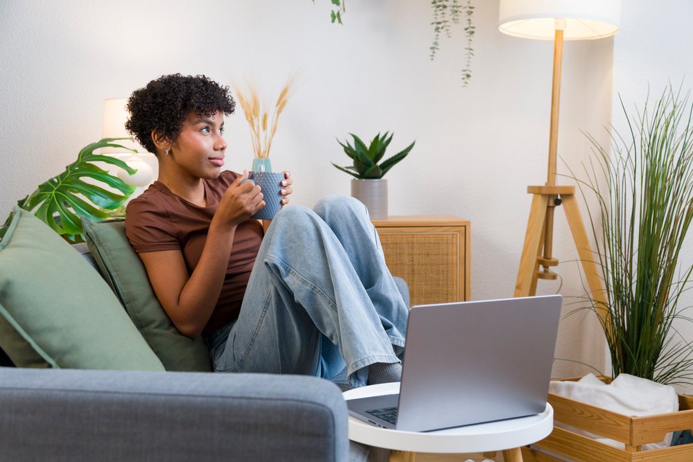 Young African American woman holding mug on sofa with laptop