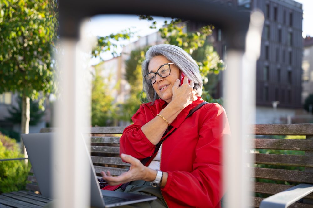 White woman in red jacket talking on phone with laptop on bench