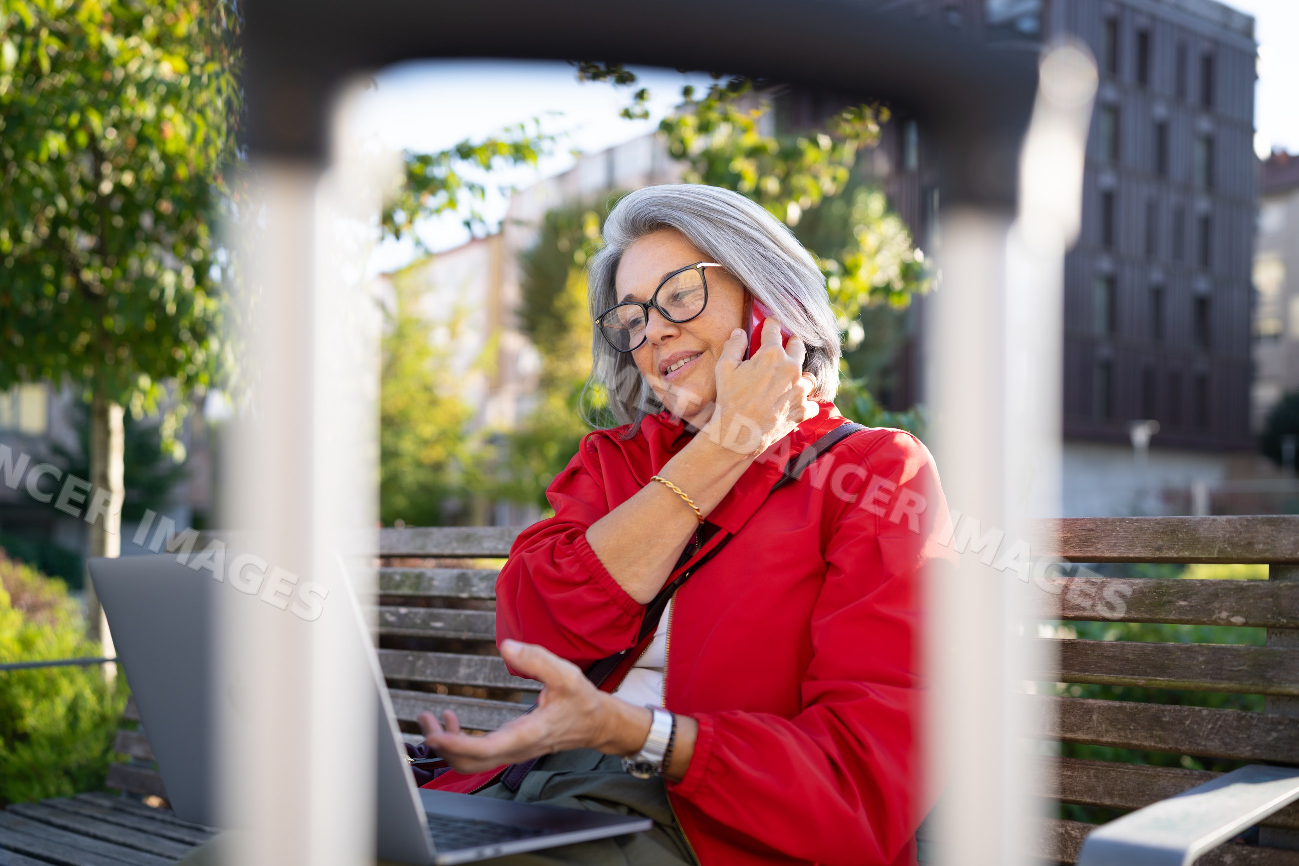 White woman in red jacket talking on phone with laptop on bench