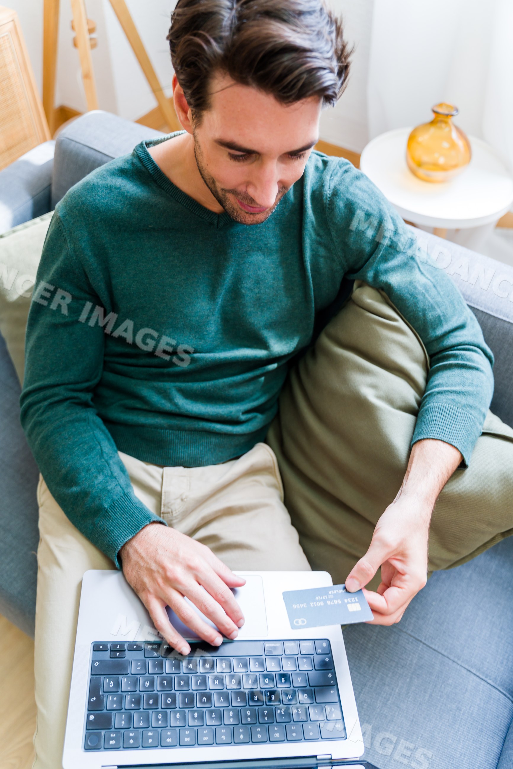 White man holding credit card using laptop on couch at home
