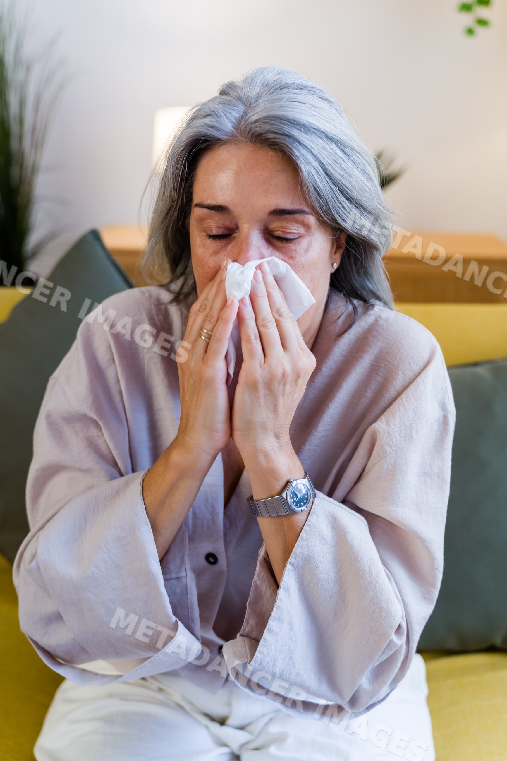 White woman blowing nose with tissue on couch indoors