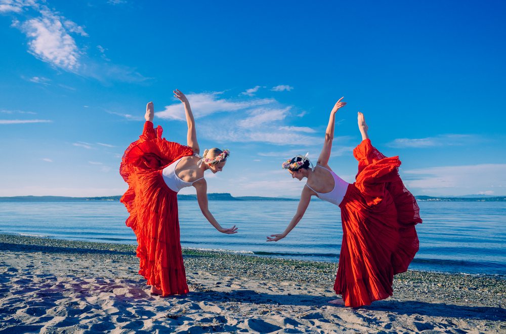 Two white women perform partner yoga acrobatics on a sandy beach