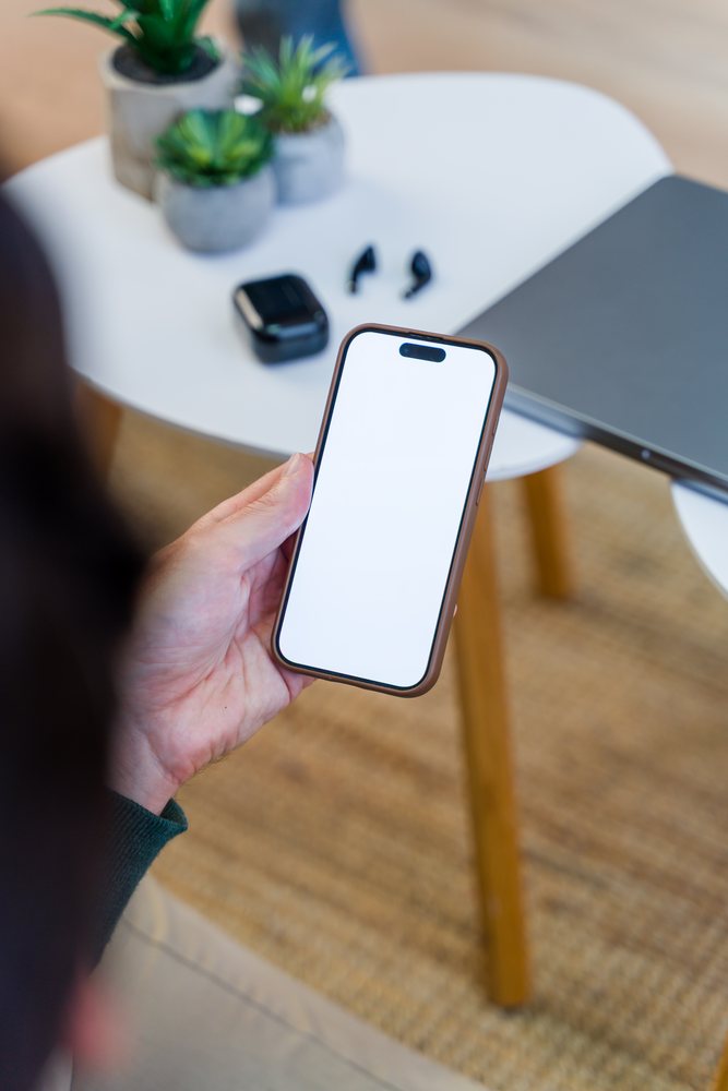 Hand holding smartphone near table with wireless earbuds and laptop