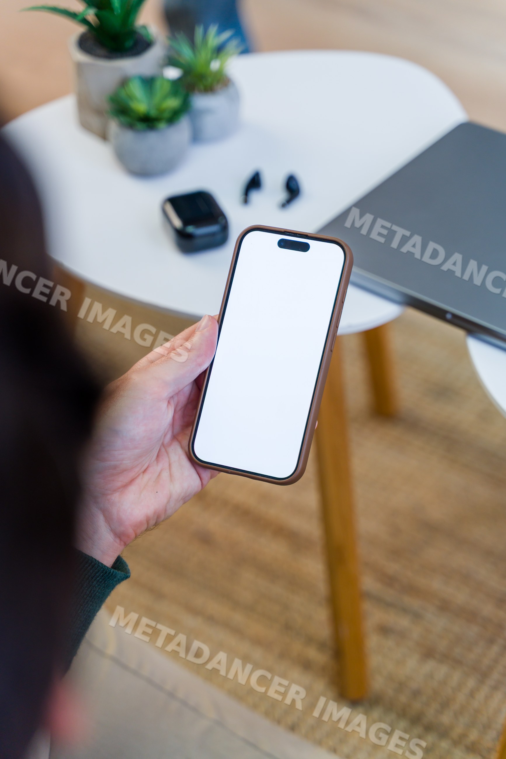 Hand holding smartphone near table with wireless earbuds and laptop