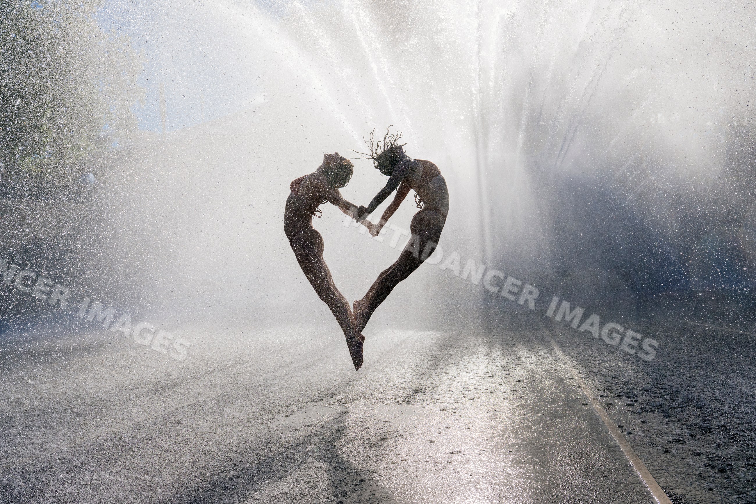 Two white women jumping hand in hand under water spray outdoors