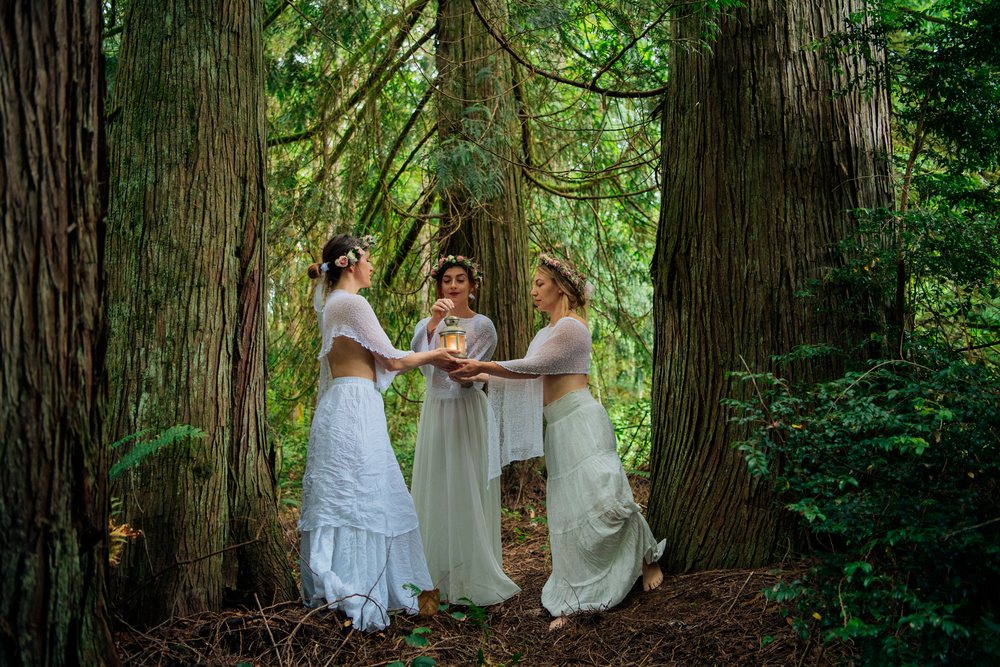 Three white women hold lantern standing among tall trees