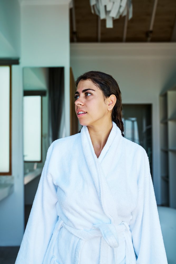 A woman wearing a white bathrobe looks thoughtfully indoors in a home