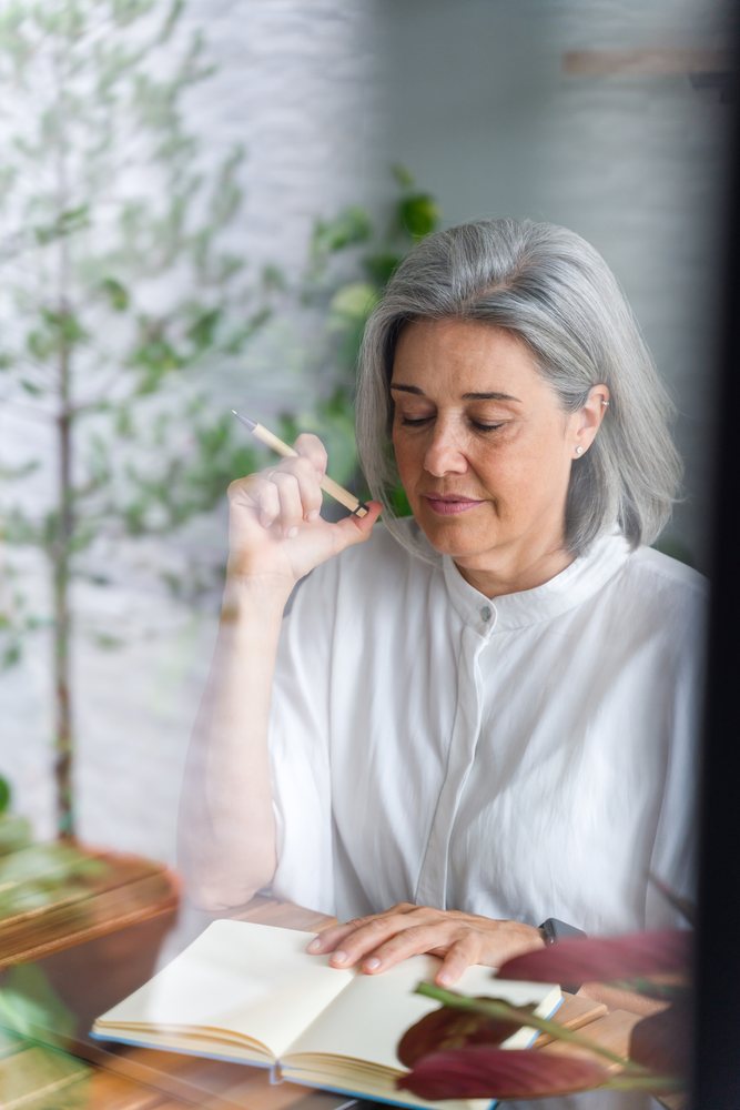 White Woman with grey hair writing in notebook indoors with plants