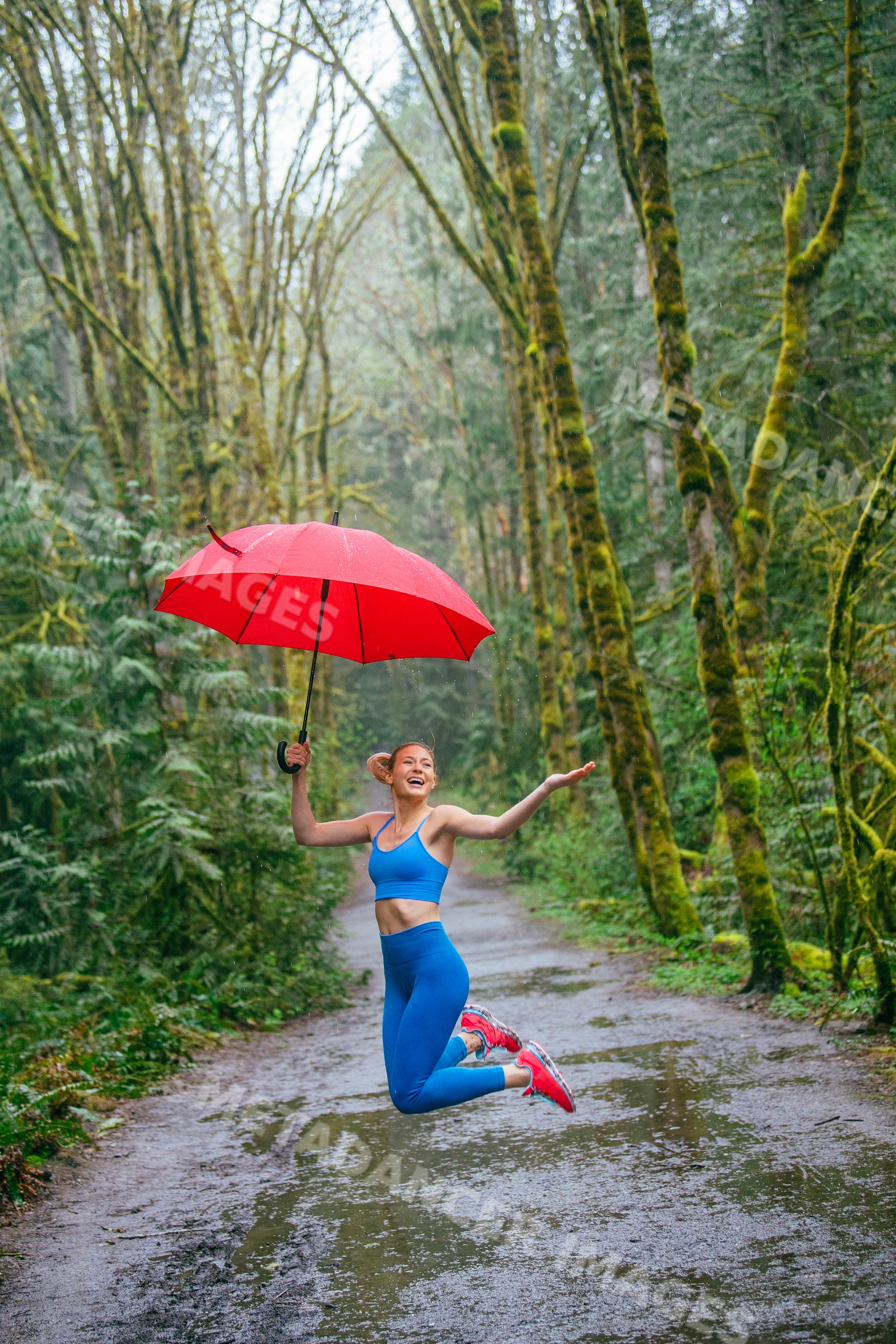 White woman jumping on wet forest trail holding red umbrella in rain