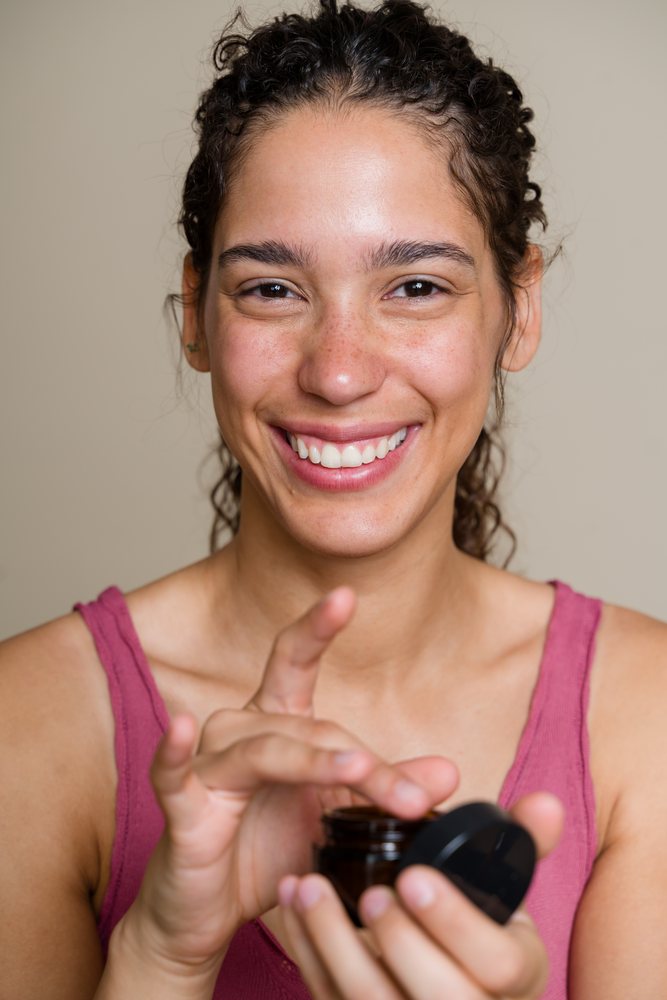 Hispanic woman applying face cream from a brown jar indoors