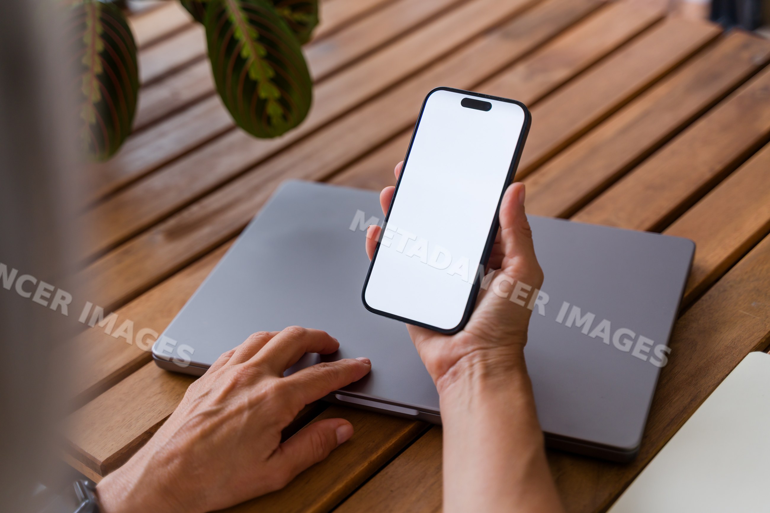 Hand holding smartphone with blank screen over laptop on wooden table