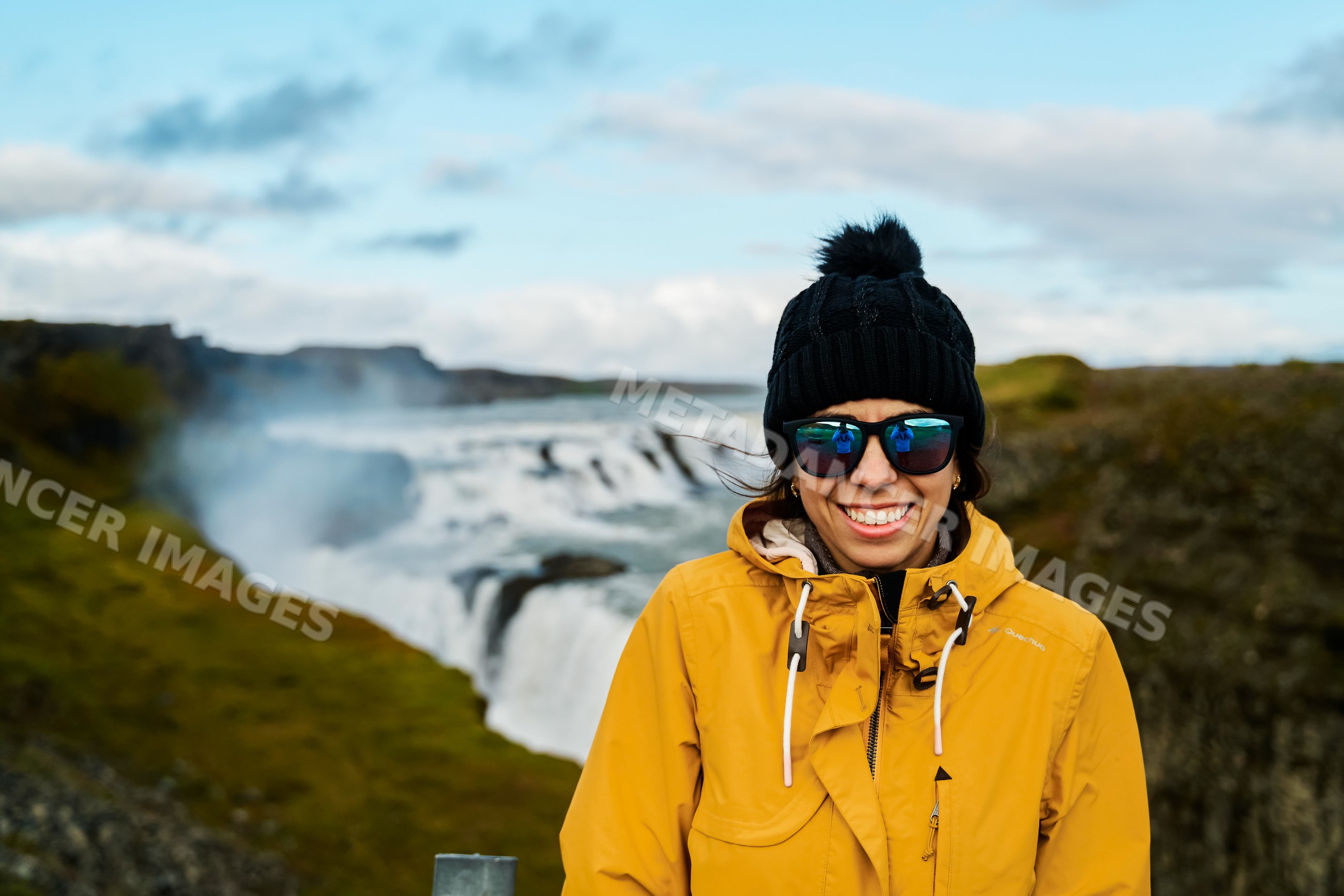 A smiling white woman in a yellow jacket stands near a waterfall