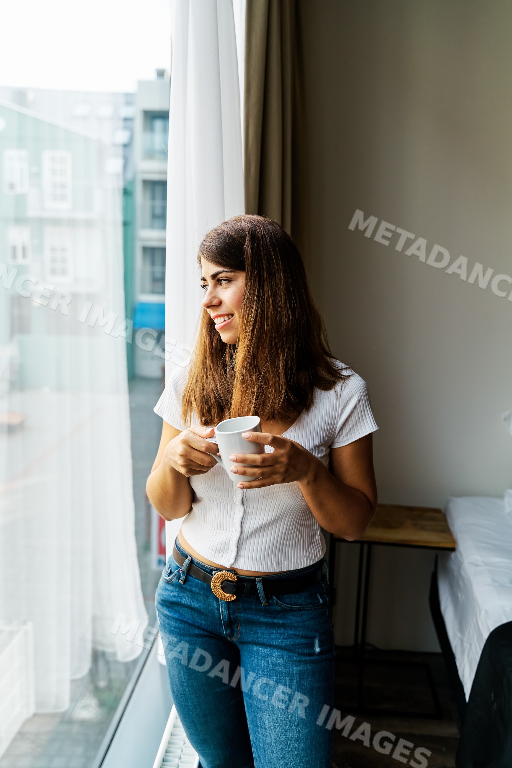 A woman holding a cup looks outside in a hotel room in a hotel