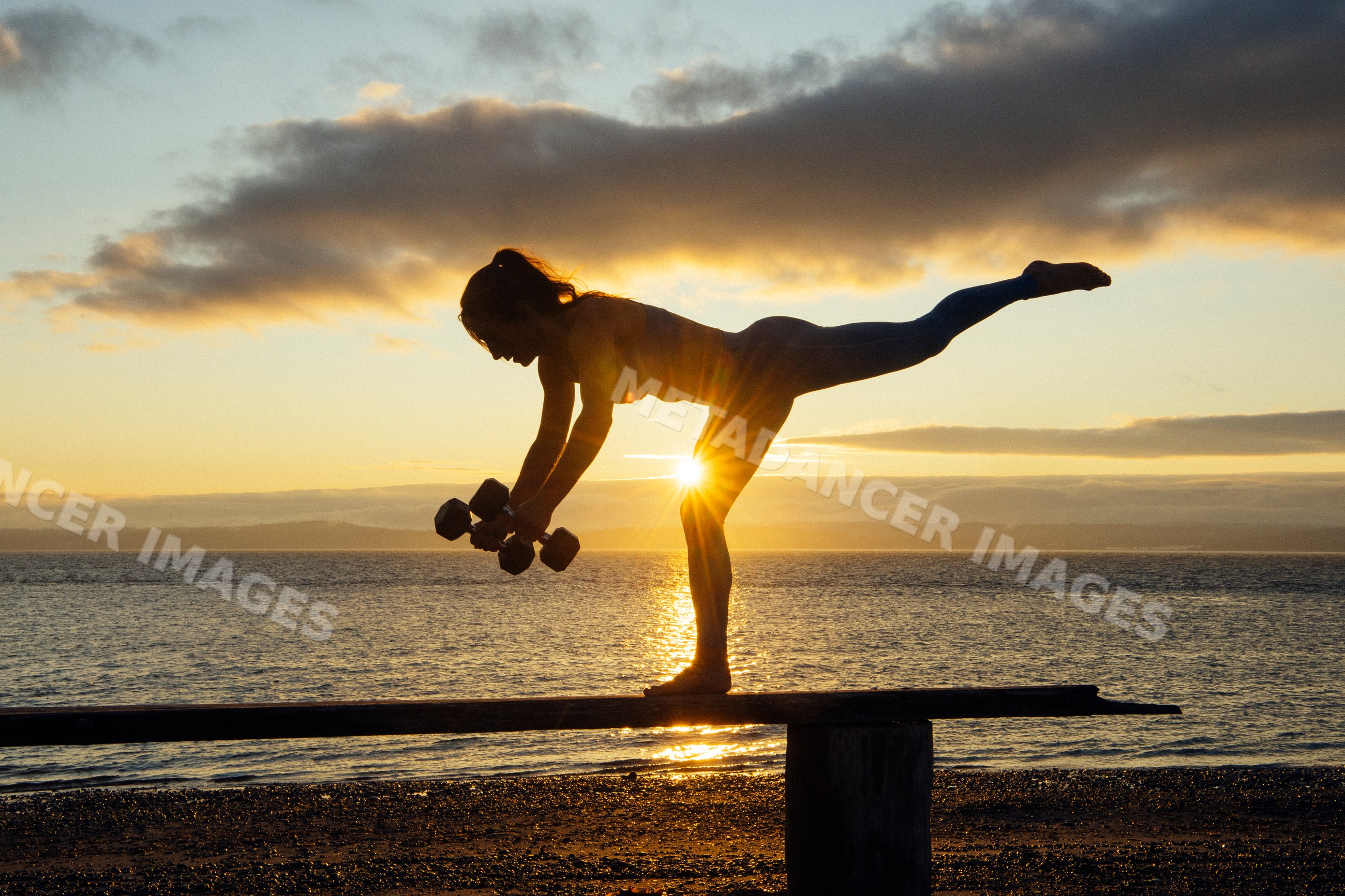 White woman exercising in balance pose with dumbbells at sunset