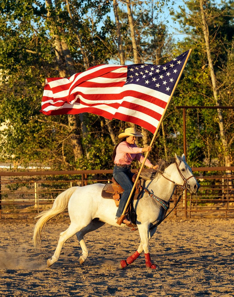 Cowgirl riding white horse carrying american flag in outdoor arena