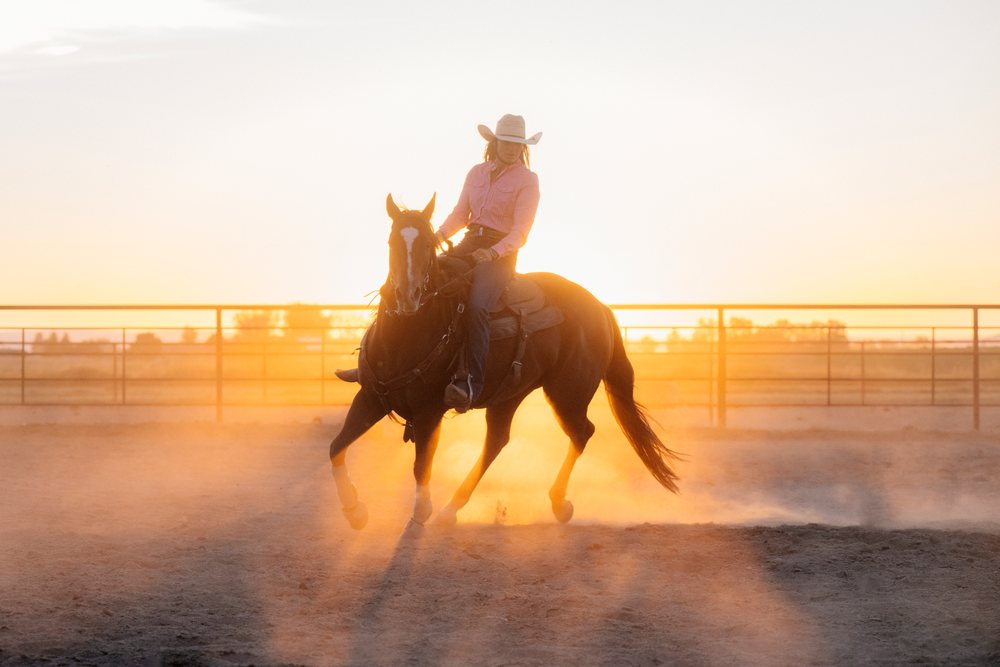 White woman riding a horse in a dusty arena at Southwest USA sunset