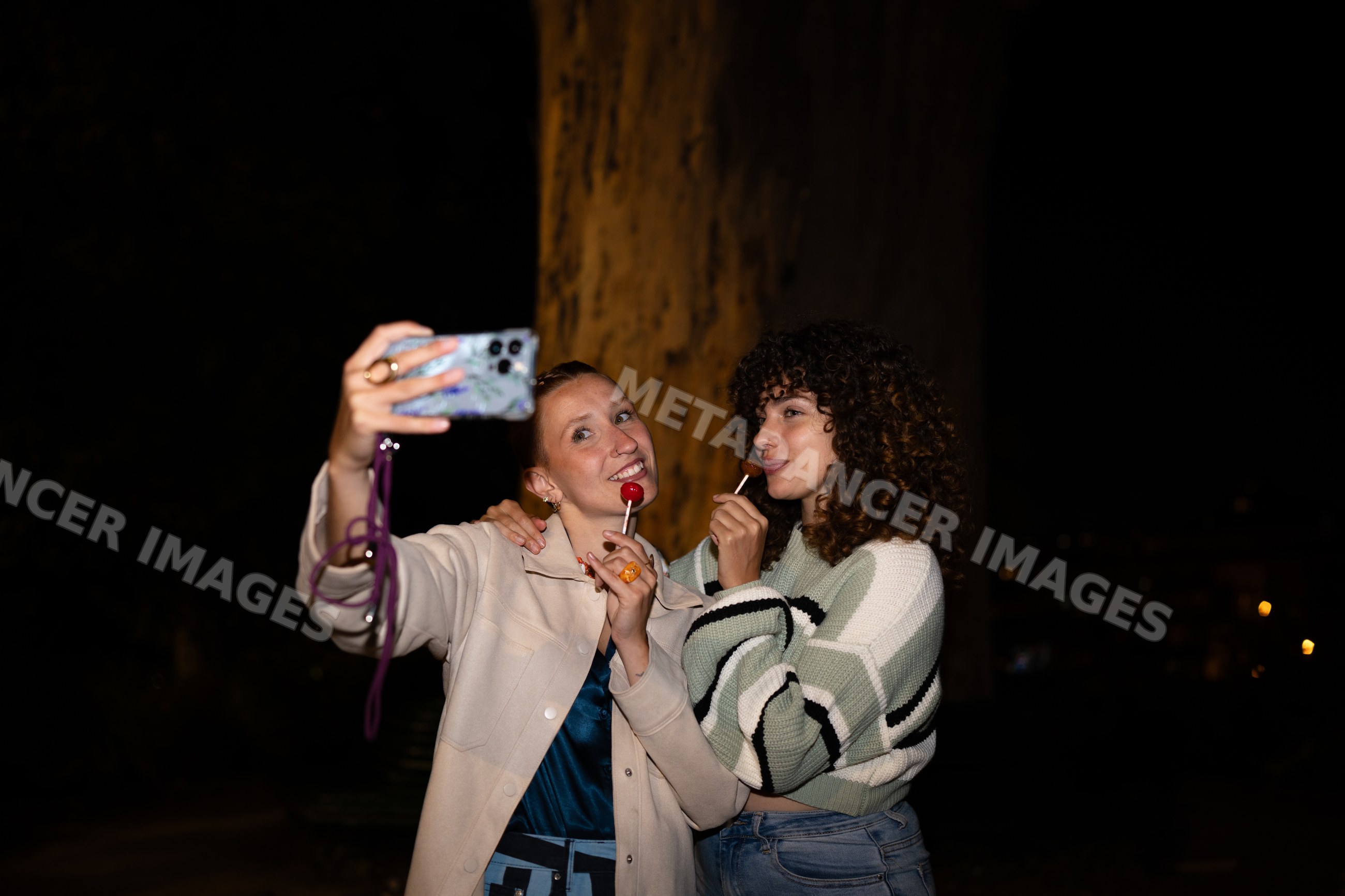 Two white women taking a selfie with lollipops outdoors at night