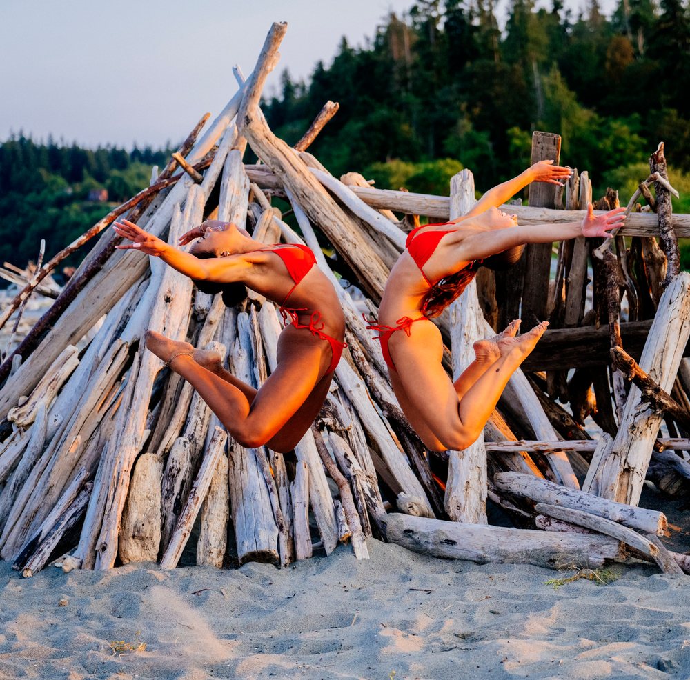 Two women jumping in red bikinis on a sandy beach with driftwood