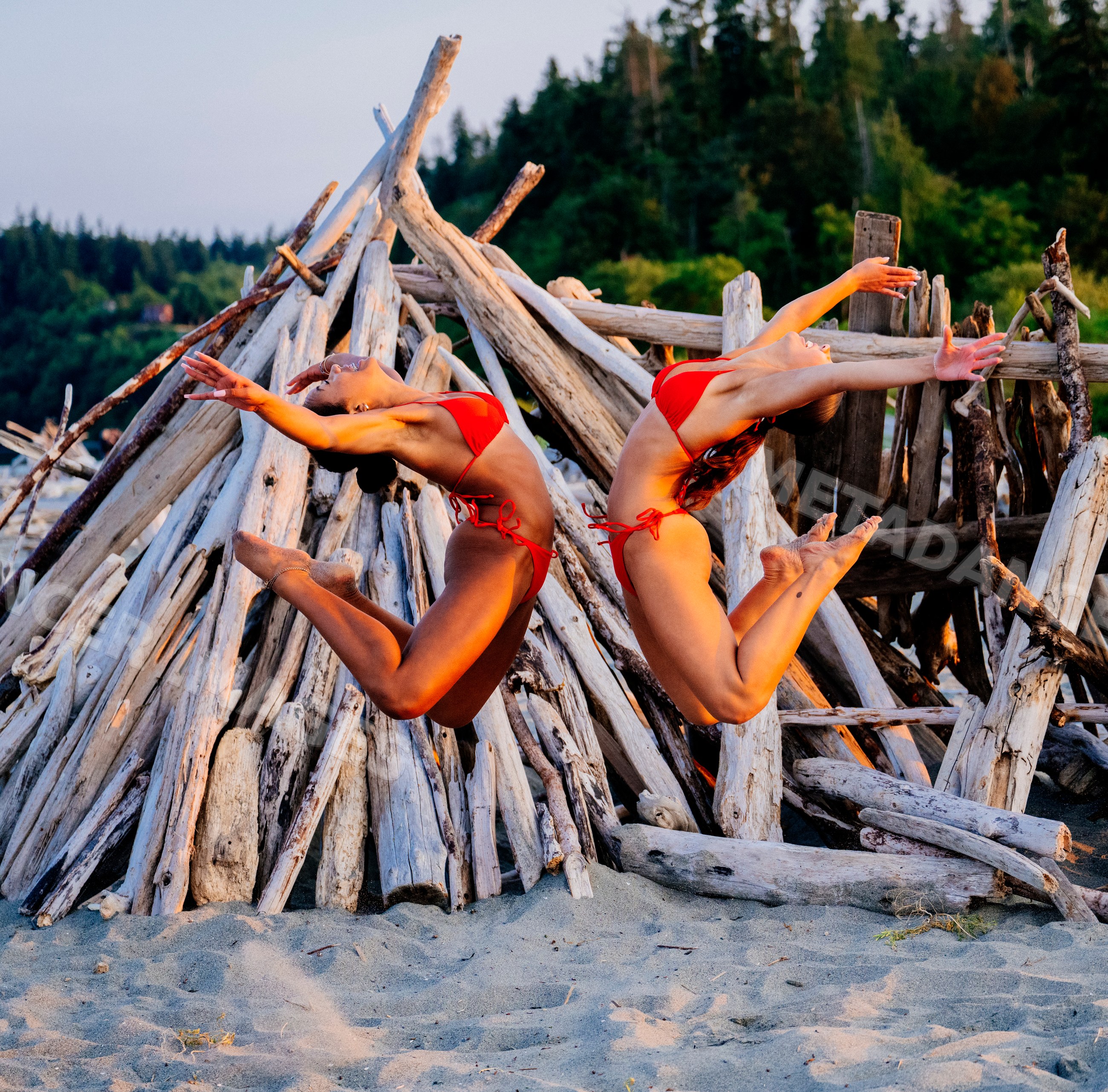 Two women jumping in red bikinis on a sandy beach with driftwood