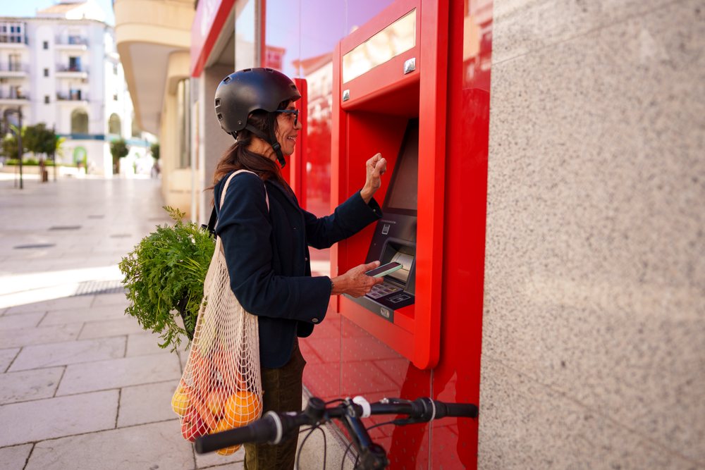 Hispanic woman using atm with bicycle and grocery bag in Málaga street