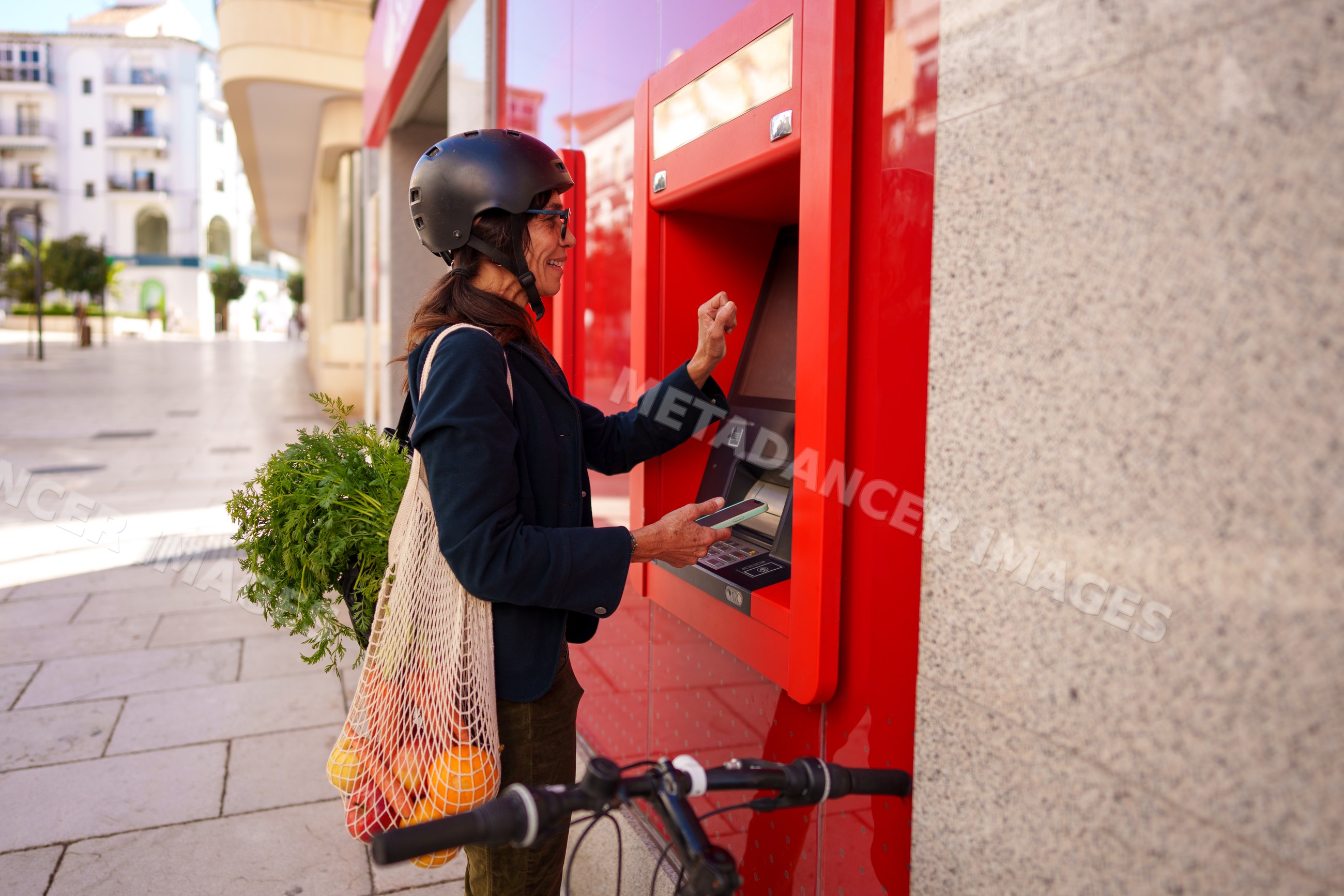 Hispanic woman using atm with bicycle and grocery bag in Málaga street